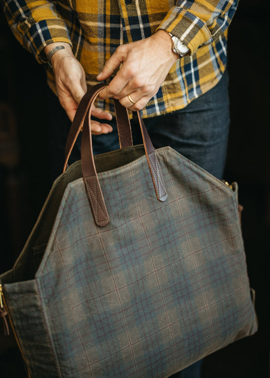 Person holding a Chilao plaid waxed canvas Newcomb tote bag with brown leather straps against a dark background