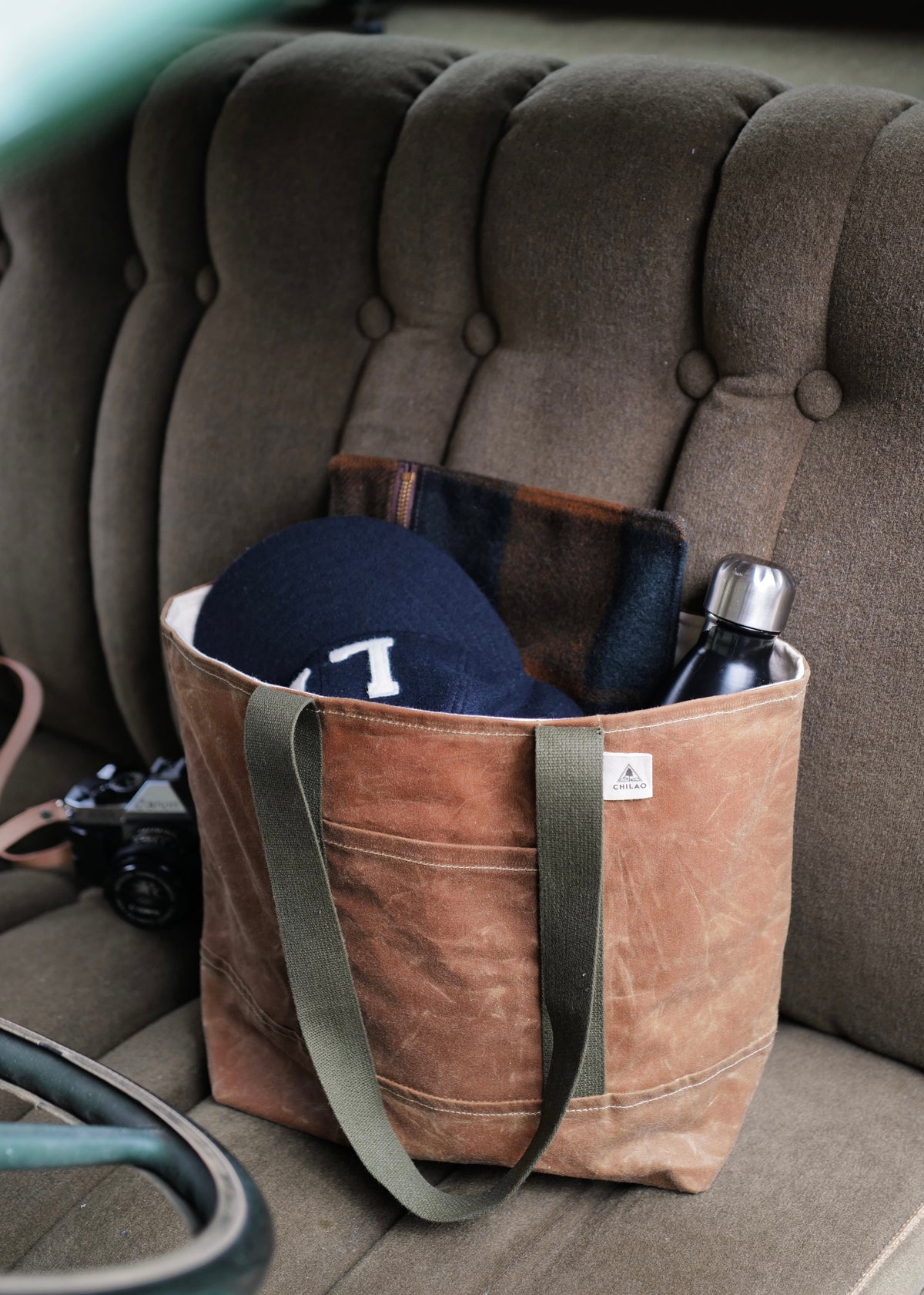 A brown waxed canvas tote bag with green military straps and a natural canvas liner, featuring two exterior pockets and one large main pocket, is displayed on a seat.