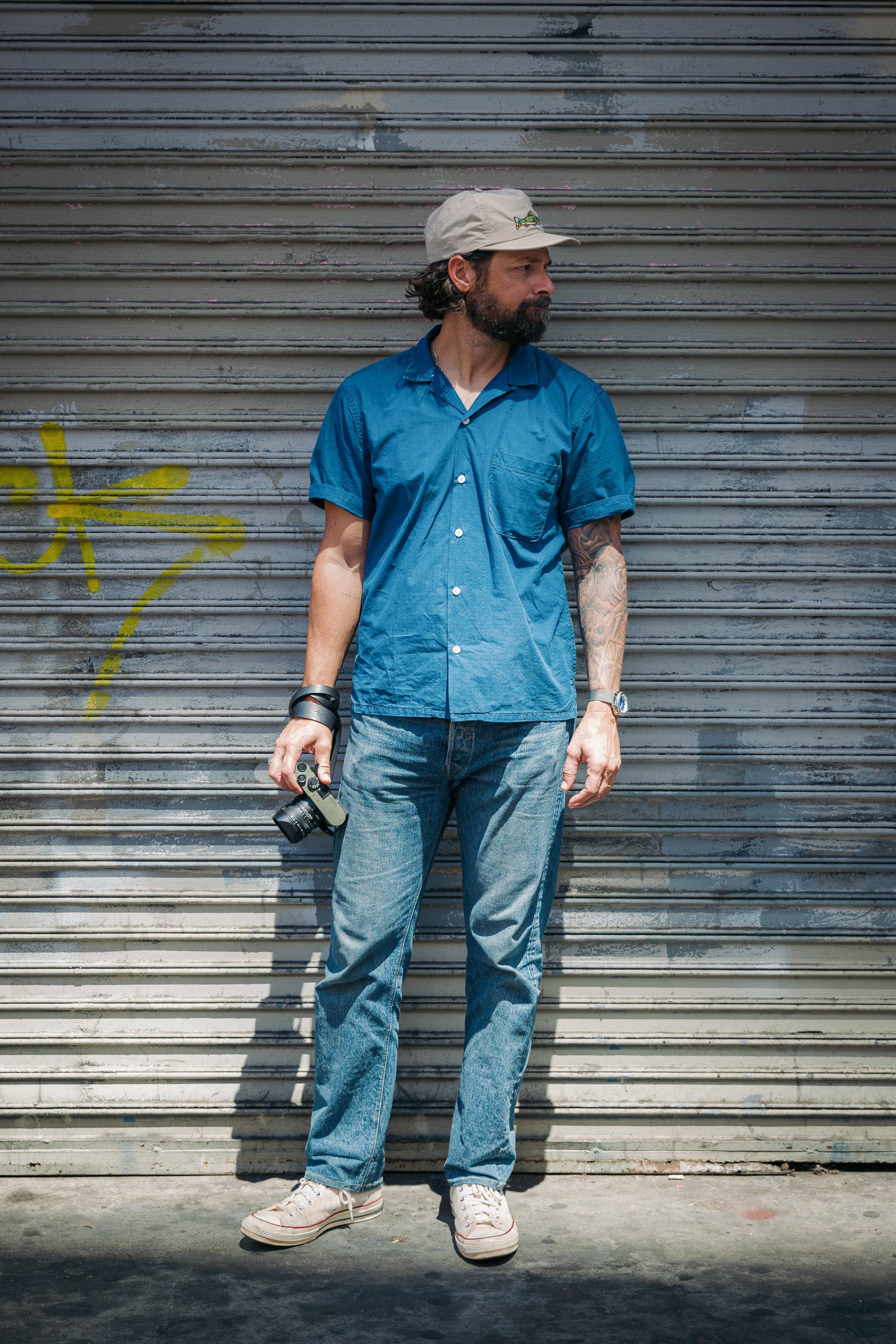 A man stands in front of a closed metal roll-up door wearing a blue short-sleeve button-up shirt, blue jeans, and beige sneakers, holding a camera.