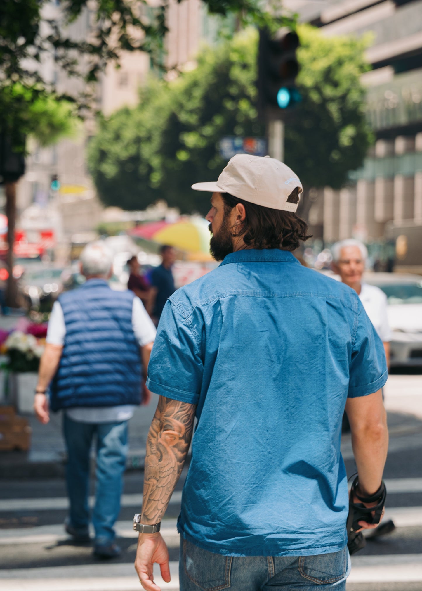 A man wearing a blue short-sleeve button-up shirt and a beige baseball cap walks across a city street crosswalk, viewed from behind.