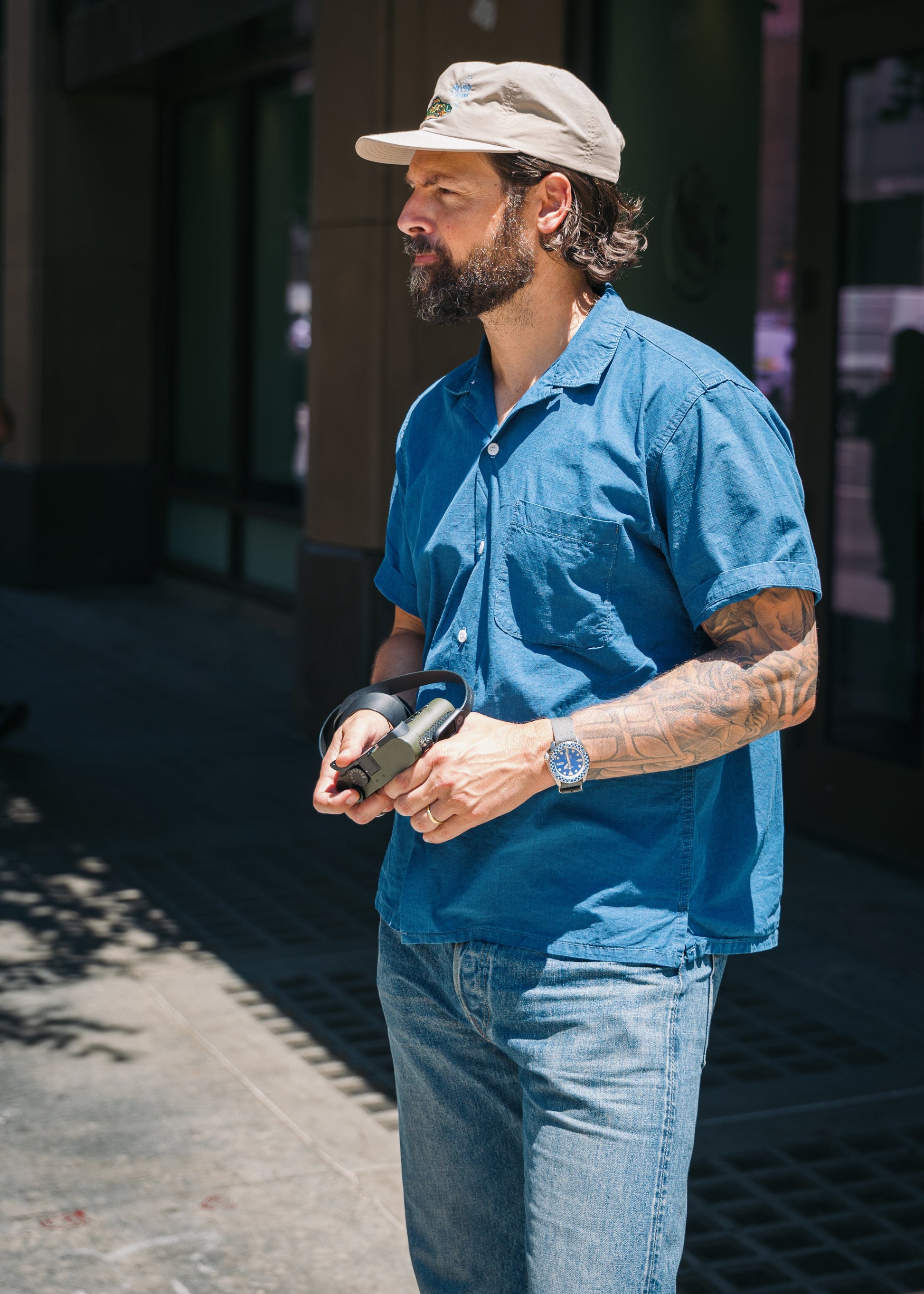 A man wearing a blue short-sleeve button-up shirt, blue jeans, and a beige baseball cap stands outdoors on a sidewalk, holding a black camera.