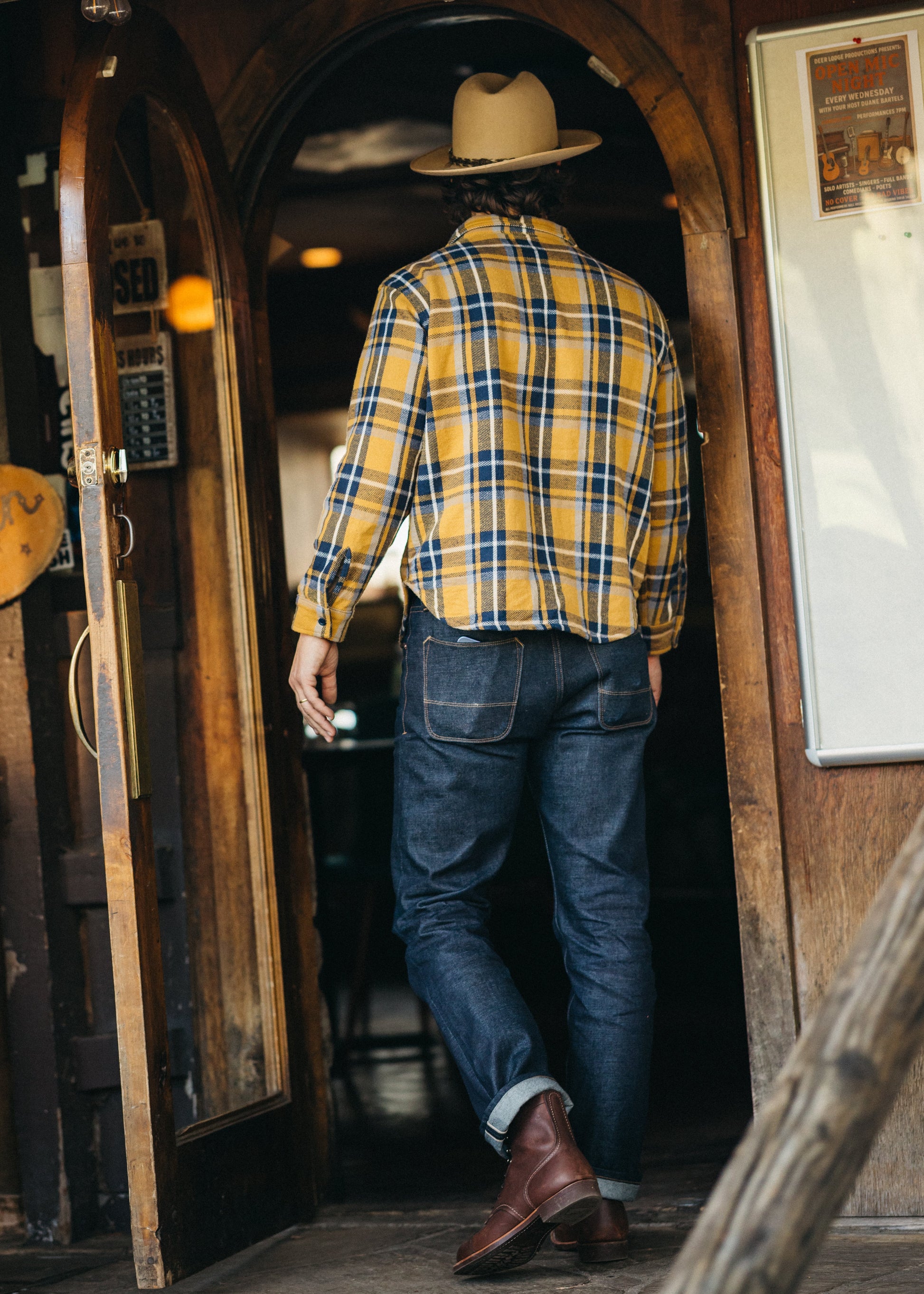 Man in a yellow plaid shirt and jeans walking through a wooden archway.