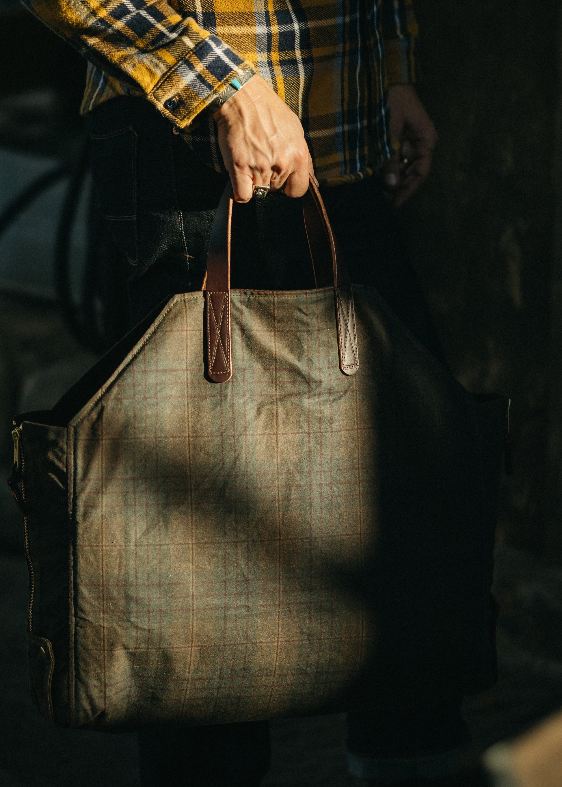 Person holding a brown leather and plaid waxed canvas bag with a dark background