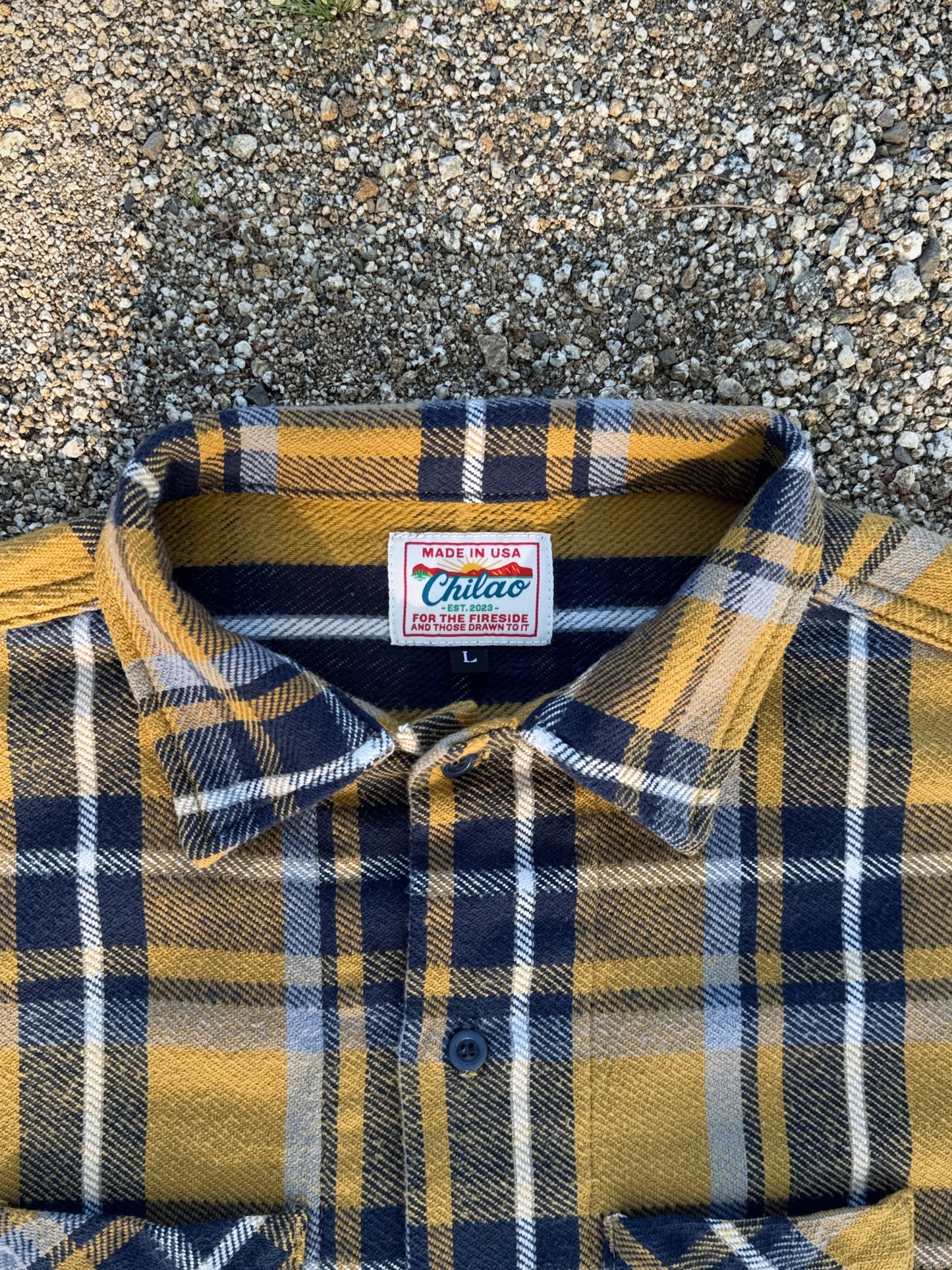 Folded plaid shirt with a Chilao brand label on a textured rocky surface. Japanese brushed twill flannel in yellow and blue. 
