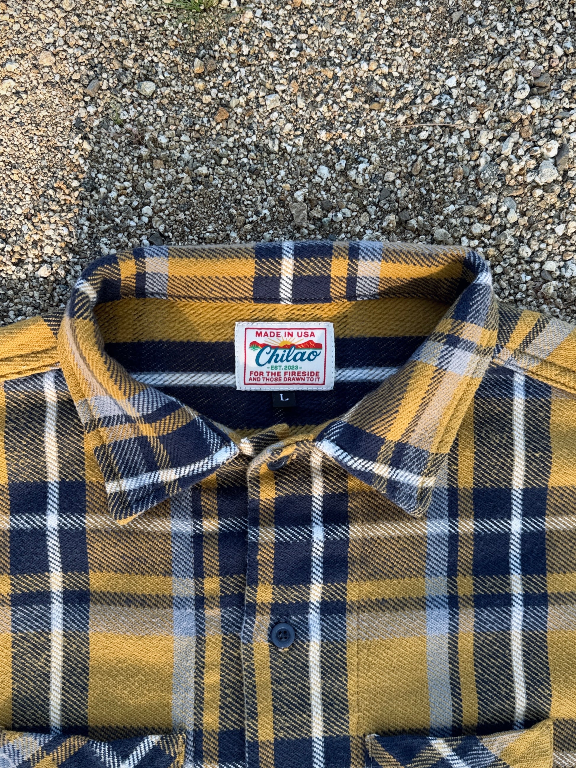 Folded plaid shirt with a Chilao brand label on a textured rocky surface. Japanese brushed twill flannel in yellow and blue. 