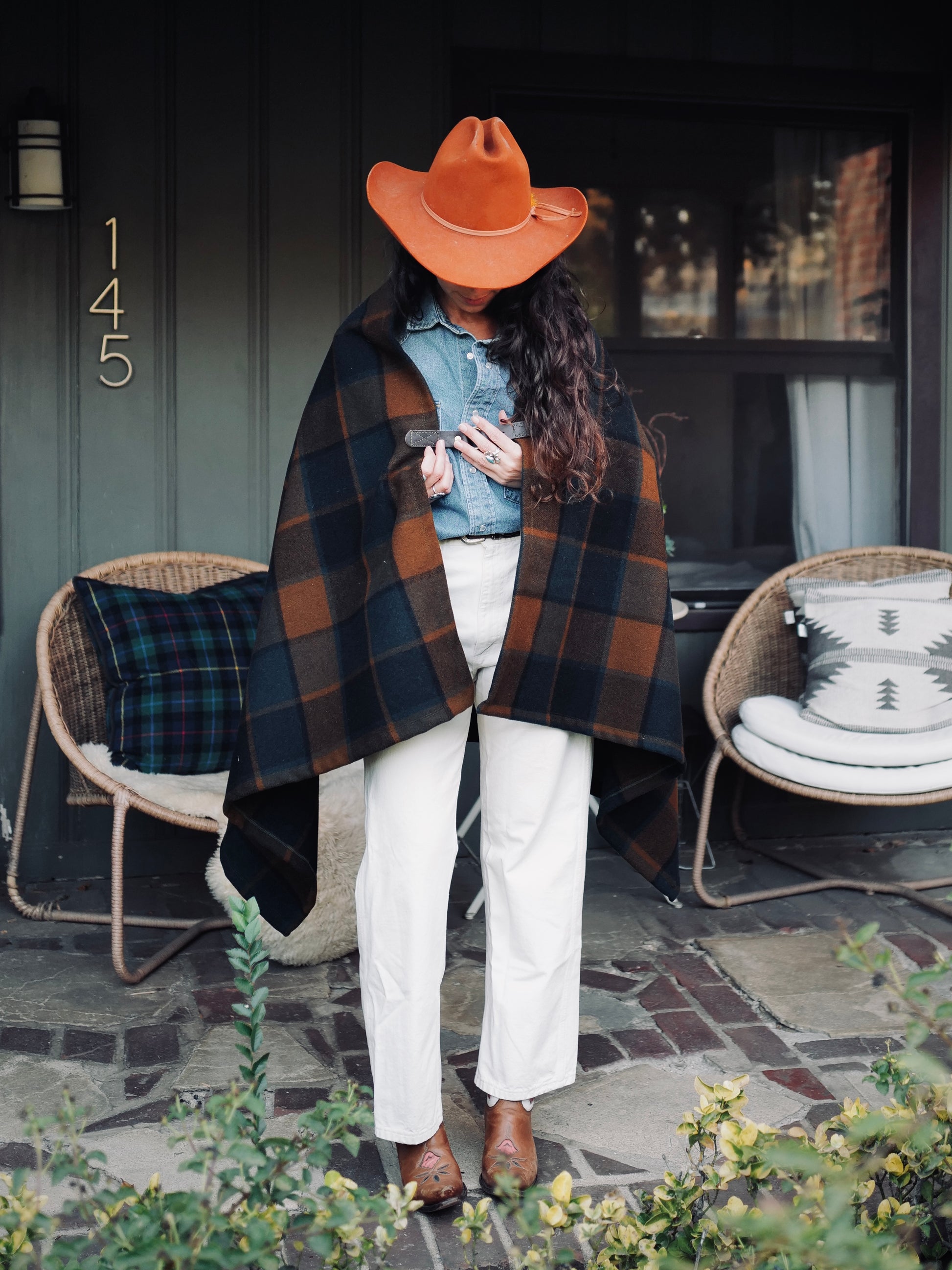 A person wearing a plaid wool poncho with leather details, paired with white pants and brown shoes, standing outdoors.