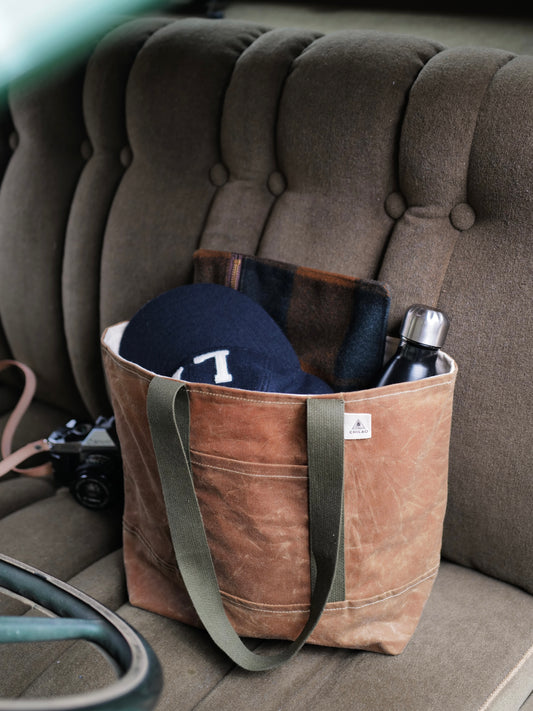 A brown waxed canvas tote bag with green military straps and a natural canvas liner, featuring two exterior pockets and one large main pocket, is displayed on a seat.